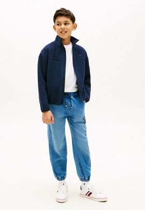 Dreng stående iført en marineblå fleecejakke, hvid t-shirt, blå joggerjeans og hvide sneakers med røde og sorte striber.