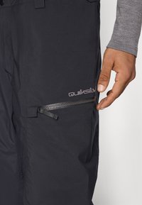 Calças pretas impermeáveis com superfície texturizada, apresentando um bolso com fecho éclair na lateral e um logótipo de cor clara próximo ao bolso.