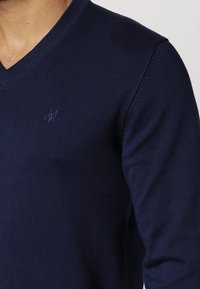 Granatowy sweter robiony na drutach z dekoltem w kształcie litery V. Posiada subtelne haftowane logo na lewej piersi oraz ściągacze na mankietach i u dołu.