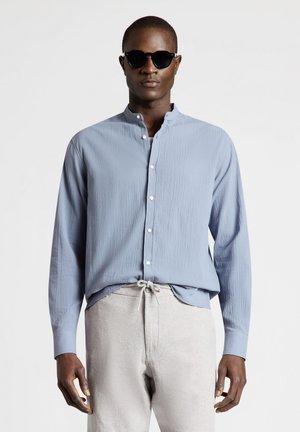 Homme portant des lunettes de soleil noires, une chemise bleu clair à manches longues avec col mandarin et un pantalon beige clair à cordon de serrage.