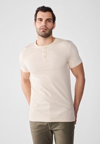 Beige kortermet T-skjorte med rund hals og knapplacket. Laget av glatt stoff, tettsittende design, kombinert med olivengrønne bukser.
