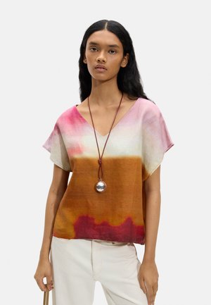 Młoda kobieta z długimi ciemnymi włosami, ubrana w bluzkę z dekoltem w serek w gradientowych kolorach różu, bieli i brązu oraz z okrągłym wisiorkiem na szyi, stojąca na jednolitym tle.