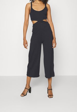 Femme portant une combinaison noire sans manches avec des découpes à la taille et un pantalon large raccourci, associée à des sandales à talons noires.