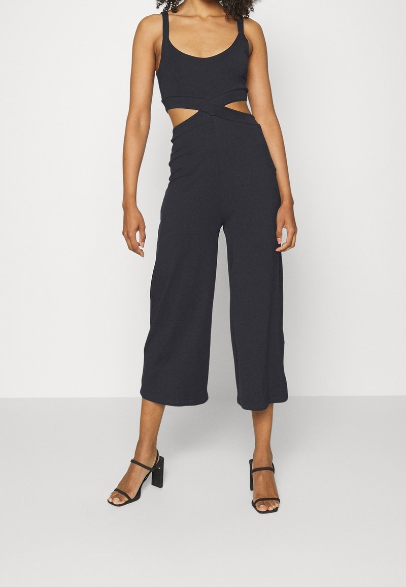 Femme portant une combinaison noire sans manches avec des découpes à la taille et un pantalon large raccourci, associée à des sandales à talons noires.
