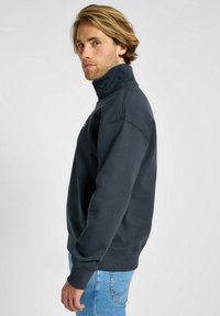 Felpa zip-up grigio scuro con colletto alto e polsini a costine, indossata con jeans azzurri chiari. Texture liscia, design minimalista.
