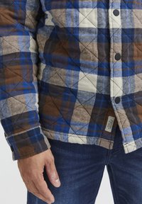 Quiltet flanellskjorte i blå, brun og kremmønstret rutete. Har svarte knapper, diamantsøm og et vevd merke på kanten.