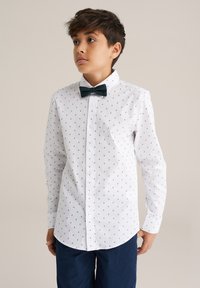 Bela srajca s gumbi in majhnimi temnimi trikotnimi vzorci, ovratnikom, dolgimi rokavi, združena s temno zeleno mašno. Vidni so temni hlače.