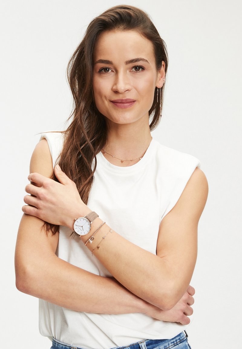 Runde Uhr mit weißem Ziffernblatt und silbernen Markierungen, graues Stoffarmband. Modell trägt geschichtete goldene Halsketten und Armbänder. Weißer Hintergrund mit Hemd.