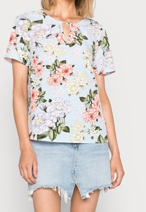 Donna che indossa un top a maniche corte floreale di colore blu chiaro con scollatura a chiave e una mini gonna in denim leggero sfilacciata con spacco frontale.