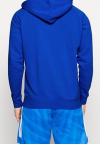 Haut en coton bleu avec un ourlet et des poignets côtelés, doté d'une capuche avec cordon de serrage. Porté avec un short bleu tie-dye avec des rayures blanches.