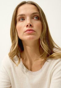 Maglione color beige chiaro con scollo ampio, morbida texture e motivo sottile. Il modello presenta lunghi capelli mossi e un'espressione neutra.