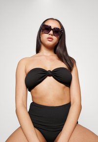 Conjunto de biquíni preto texturizado, apresentando um topo sem alças com um detalhe de nó e parte de baixo de cintura alta. Modelo usando óculos de sol oversized.