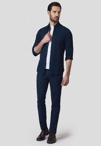 Granatowa koszula z guzikami noszona na białej koszulce, w zestawieniu z granatowymi spodniami i brązowymi butami. Materiał wydaje się gładki, a krój jest dopasowany.