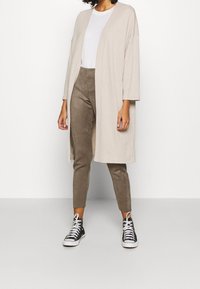 Un cardigan long beige, à l'avant ouvert, au fit décontracté, associé à un t-shirt gris clair et un pantalon slim taupe. Des baskets montantes noires complètent le look.