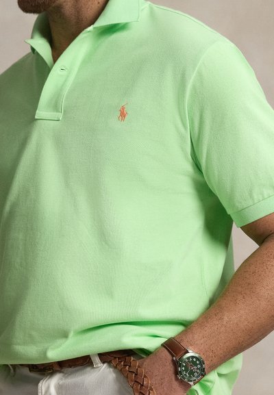 Ανοιχτό πράσινο πόλο με υφή, με ένα μικρό πορτοκαλί κεντημένο λογότυπο. Φορεμένο με καφέ πλεκτή ζώνη και πράσινο ρολόι.