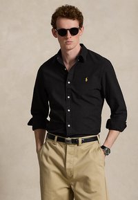 Bărbat care poartă o cămașă neagră cu nasturi și logo galben, pantaloni bej, curea neagră, ochelari de soare închiși la culoare și ceas de mână, stând cu mâinile în buzunare.