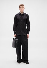 Fekete hosszú ujjú ing és hozzáillő, bő szárú nadrág. A modell egy fekete bőr táskát tart, amelynek felső fogói vannak és sima felületű.