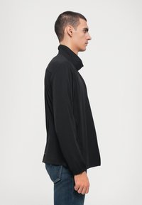 Jaqueta preta leve com colarinho alto, mangas longas e um bolso com fecho éclair. Feita de material suave e flexível, com um design minimalista.