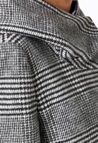 Lähikuva henkilöstä, joka pukeutunut mustavalkoiseen saksanpähkinäkuvioituun villakangastakkiin, jossa on suuri, taiteltu kaulus.