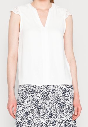 Haut blanc sans manches avec manches en dentelle à œillets, associé à une jupe à motifs tachetés noirs et blancs sur une personne debout.