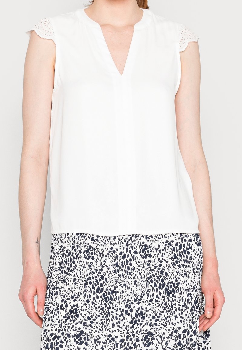 Haut blanc sans manches avec manches en dentelle à œillets, associé à une jupe à motifs tachetés noirs et blancs sur une personne debout.