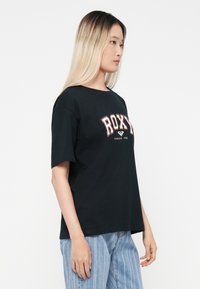 Musta puuvillane T-särk, millel on julge "ROXY" logo punases ja valges, lühikesed varrukad ja lahtine lõige. Kombineeritud helesiniste triibuliste teksadega.