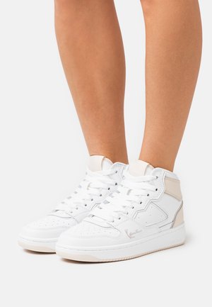 Zapatillas blancas de caña alta con detalles beige, usadas sobre piernas desnudas contra un fondo blanco liso.