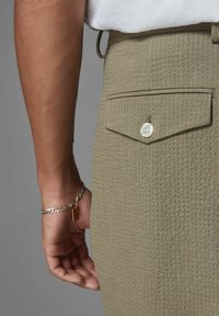 Olive green textured trousers with a back pocket featuring a white button, accompanied by a silver chain bracelet on the wrist.