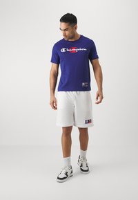 Camiseta de algodón azul con el logo de "Champion", combinada con pantalones cortos de malla blanca con bolsillos laterales y parche de la marca, calcetines blancos y zapatillas deportivas.