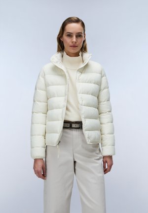 Frau trägt eine weiße Steppjacke über einem cremefarbenen Rollkragenpullover und hellbeigen Hosen und steht vor einem einfarbigen hellen Hintergrund.