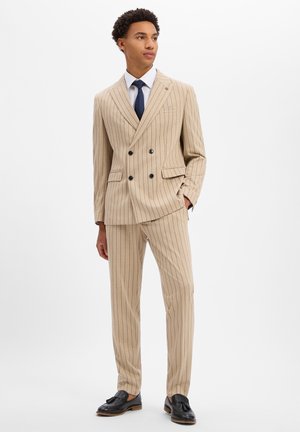 Jonge man in beige krijtstreep dubbelgeknoopt pak met wit overhemd, donkerblauwe stropdas en zwarte loafers met kwastjes, staand met één hand in de zak.