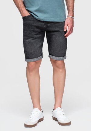 Mann trägt hochgekrempelte schwarze Jeansshorts, ein hellblaues Hemd, weiße Sneakers und ein Perlenarmband, steht mit einer Hand in der Tasche.
