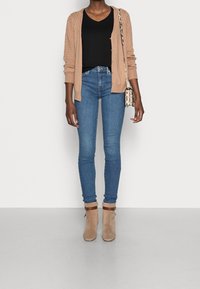 Cárdigan de punto beige sobre una blusa negra, combinado con jeans ajustados azules y botines tobilleros topo con correas marrones. Pequeño bolso cruzado con patrones.