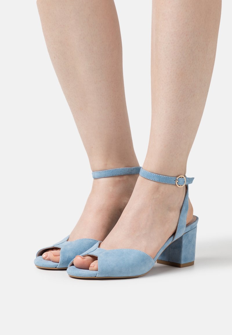 Pieds chaussés de sandales à talons blocs ouvert à l'avant en daim bleu clair avec brides à la cheville et boucle dorée sur un fond neutre.