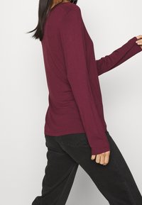 Burgunderfarbene Langarm-T-Shirts aus weichem Material, mit Rundhalsausschnitt und enger geschnittenen Ärmeln, kombiniert mit schwarzen Hosen.