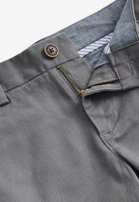 Pantalones grises con cremallera parcialmente abierta y cierre de botón, mostrando el forro interior de la pretina a rayas y costura detallada.