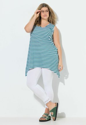 Kobieta nosząca oversize turkusowo-białą, paskowaną, bezrękawną tunikę, białe legginsy z koronkowymi mankietami, zielone sandały i okulary, stojąca przy białej ścianie.