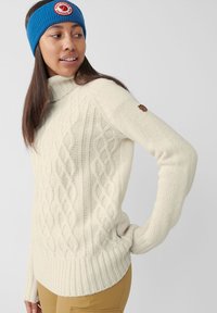 Maglione beige a trecce con colletto alto e orlo a coste. Patch con logo marrone ben visibile sulla manica e accessorio fascia in maglia blu.