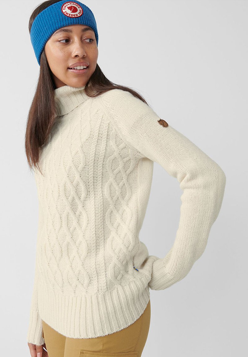 Maglione beige a trecce con colletto alto e orlo a coste. Patch con logo marrone ben visibile sulla manica e accessorio fascia in maglia blu.