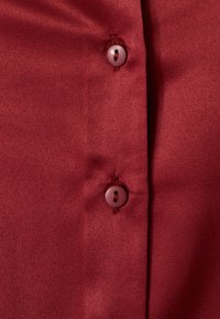 Camisa de satén rojo con una textura suave y un acabado brillante, con botones en contraste abrochados a lo largo de la tapeta delantera.