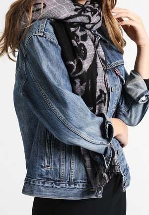 Femme portant une veste en denim bleu et une écharpe à motifs noir et gris, les bras croisés et la main près du visage.