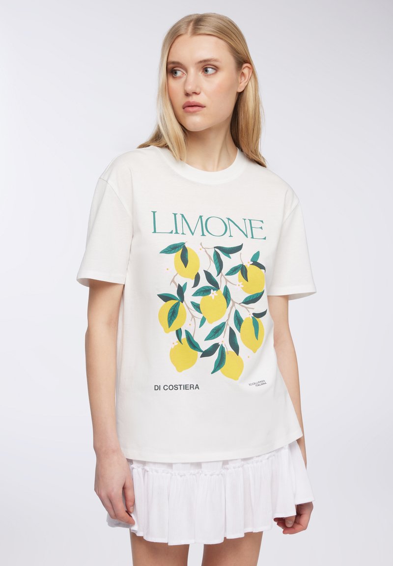 T-shirt bianca a maniche corte con stampa di limoni e foglie, con testo "LIMONE" in verde. Cotone morbido con vestibilità rilassata.