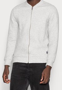 Sweat-shirt gris clair à fermeture éclair avec un motif texturé, des poignets et un ourlet côtelés, et une fermeture éclair lisse. Associé à un jean foncé.