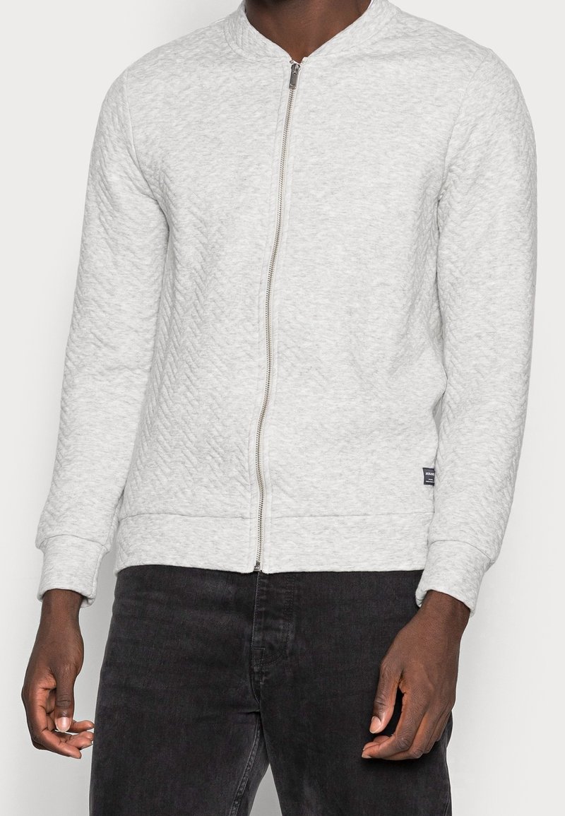 Sweat-shirt gris clair à fermeture éclair avec un motif texturé, des poignets et un ourlet côtelés, et une fermeture éclair lisse. Associé à un jean foncé.