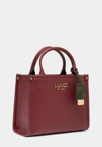 Sac à main en cuir bourgogne avec deux poignées supérieures, logo doré et une étiquette décorative. Forme carrée avec une finition texturée et des accents dorés.