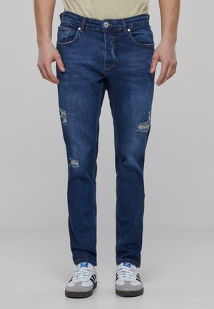 Tamne traperice u tamnoplavoj boji sa slim fit krojem, s istrošenim zakrpama na prednjoj strani i standardnim dizajnom s pet džepova.