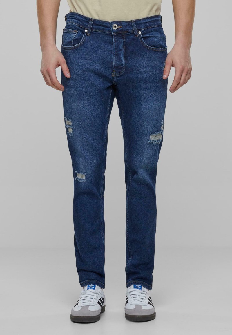 Džinsne hlače temno modre barve s slim fit krojem, ki imajo na sprednji strani obrabljene madeže in standardno pet žepno zasnovo.