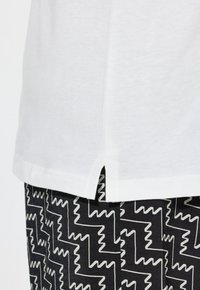 Bílá bavlněná košile s bočními rozparky, zkombinovaná s černými kalhotami s bílým zigzagovým vzorem. Měkká textura a ležérní design.