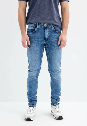 Vaqueros ajustados de mezclilla azul con un diseño clásico de cinco bolsillos, textura ligeramente desteñida y costuras en contraste. Combinados con zapatillas blancas.