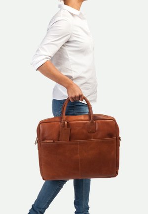 Sac à ordinateur en cuir marron avec double poignées, poche avant et étiquette de bagage. Présente une texture lisse et une forme structurée.